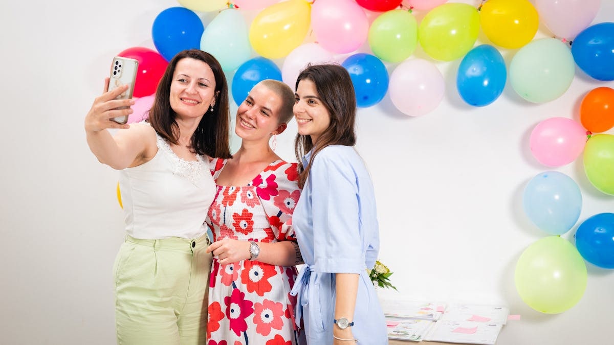 Three women smiling and taking a selfie in front of colorful balloons at a party.
